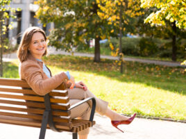 Sesja wizerunkowa ekspertki, uśmiechnięta kobieta w stroju biznesowej siedząca na ławce, w słoneczny dzień, w tle kolorowe jesienne drzewa