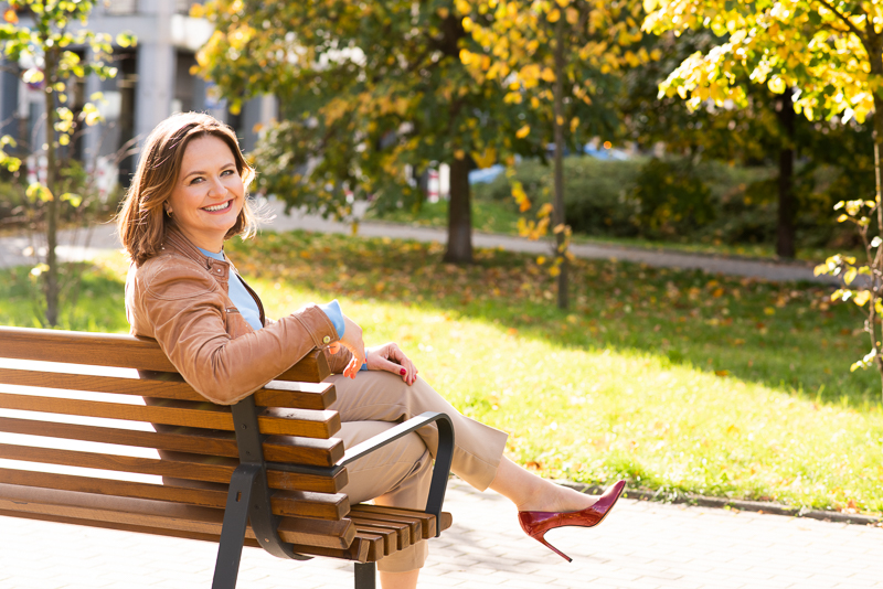 Sesja wizerunkowa ekspertki, uśmiechnięta kobieta w stroju biznesowej siedząca na ławce, w słoneczny dzień, w tle kolorowe jesienne drzewa